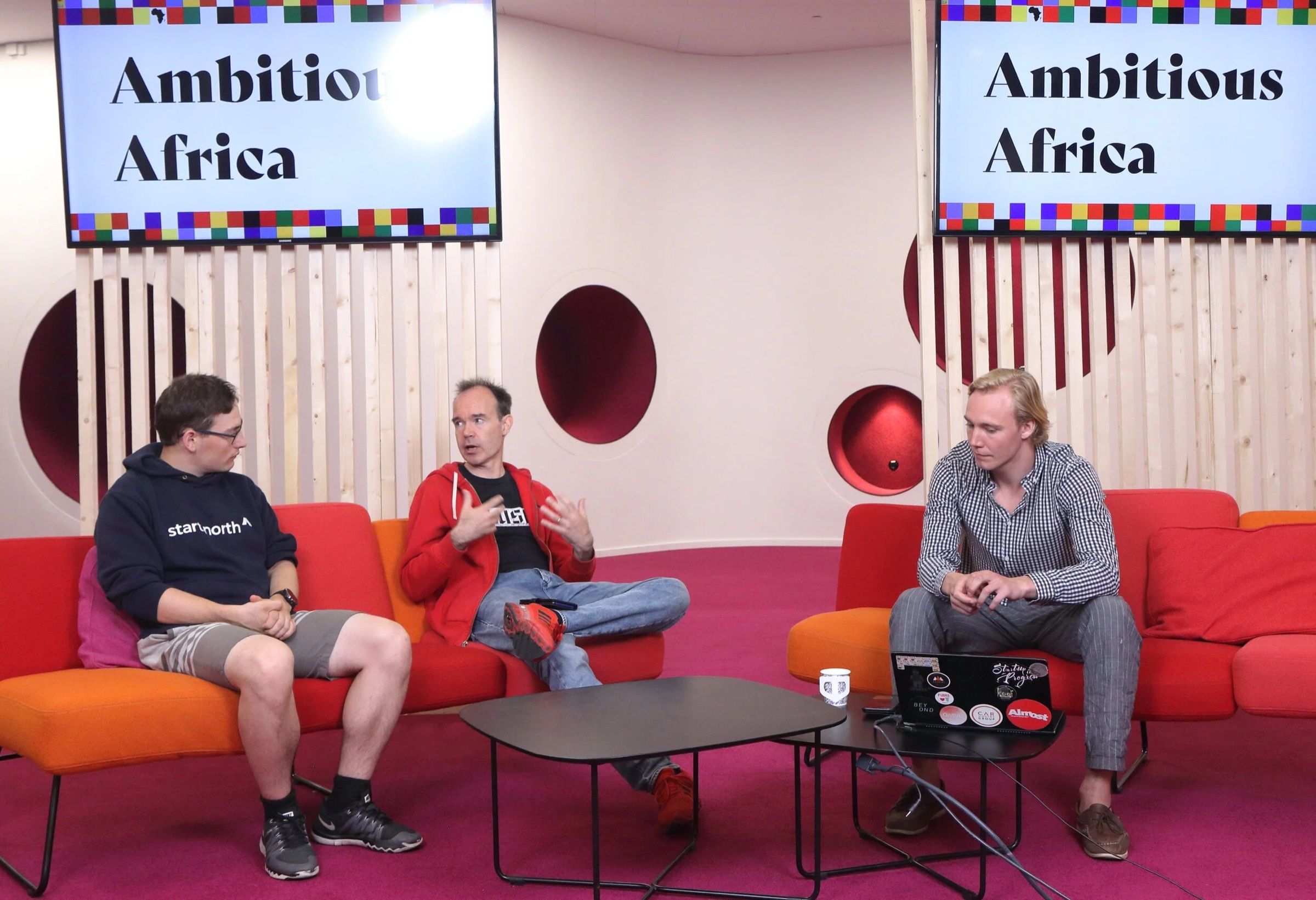 Three people on red sofas in a broadcast studio under Ambitious Africa signs