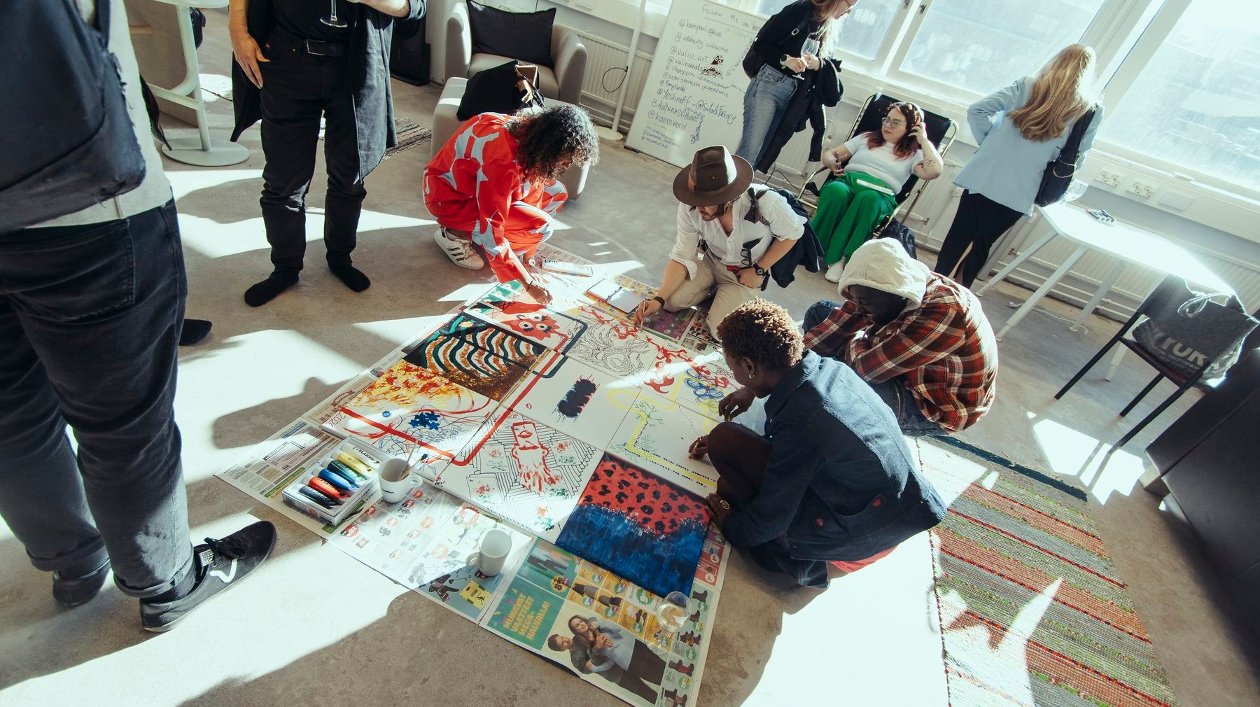 People kneeling and standing around large sheets of paper spread across a sunlit floor, painting together