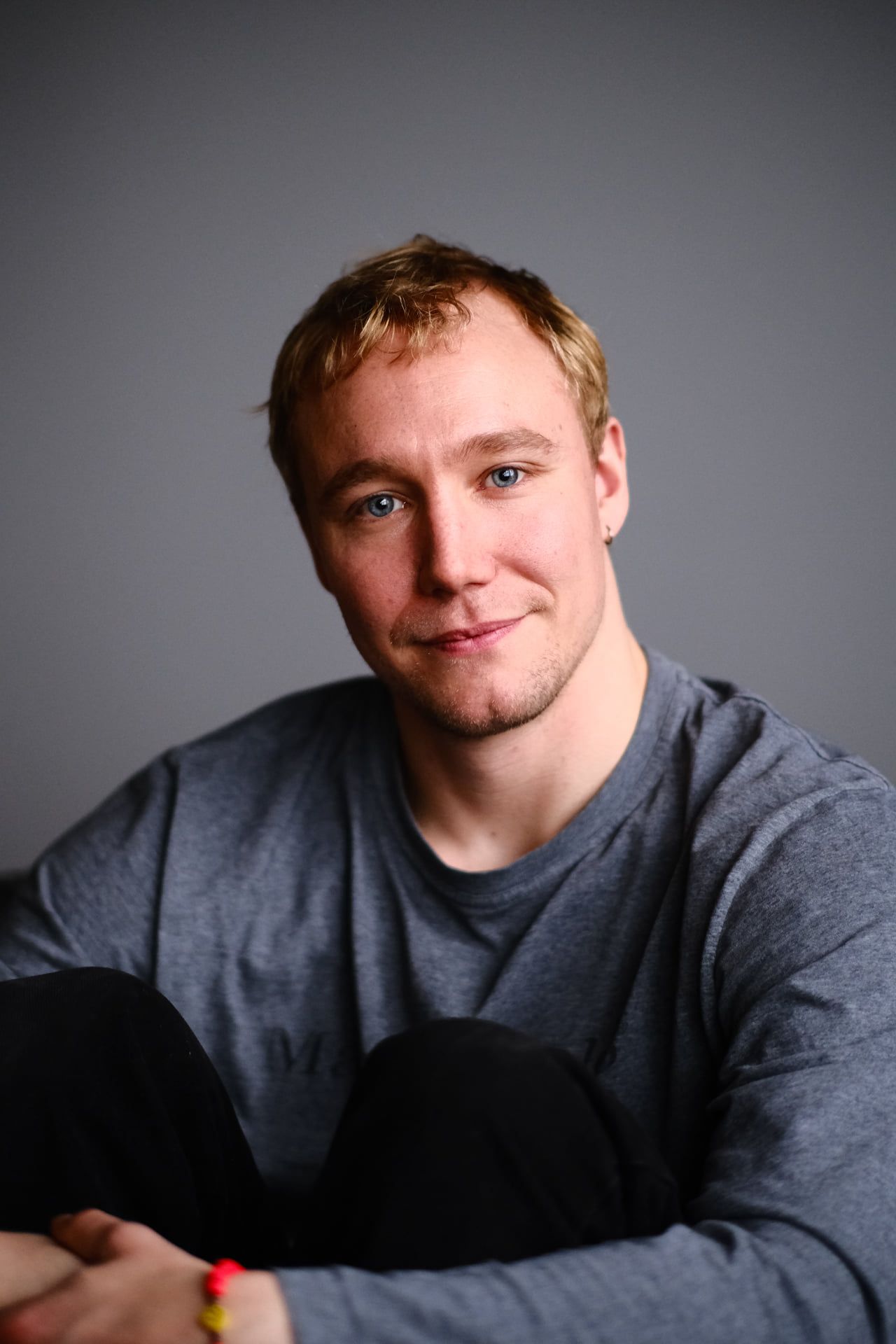 Ronny Eriksson seated on a grey studio floor, arms around his knees, smiling slightly at the camera