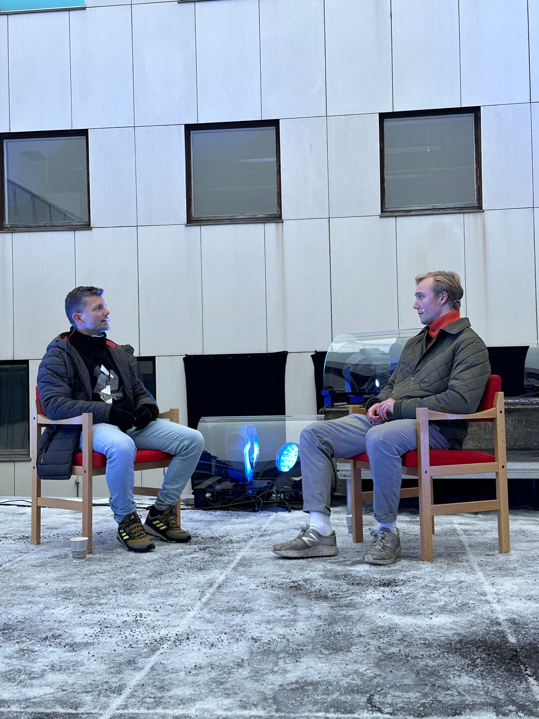 Two men in chairs on an outdoor stage in Kirkenes, a fire bowl between them