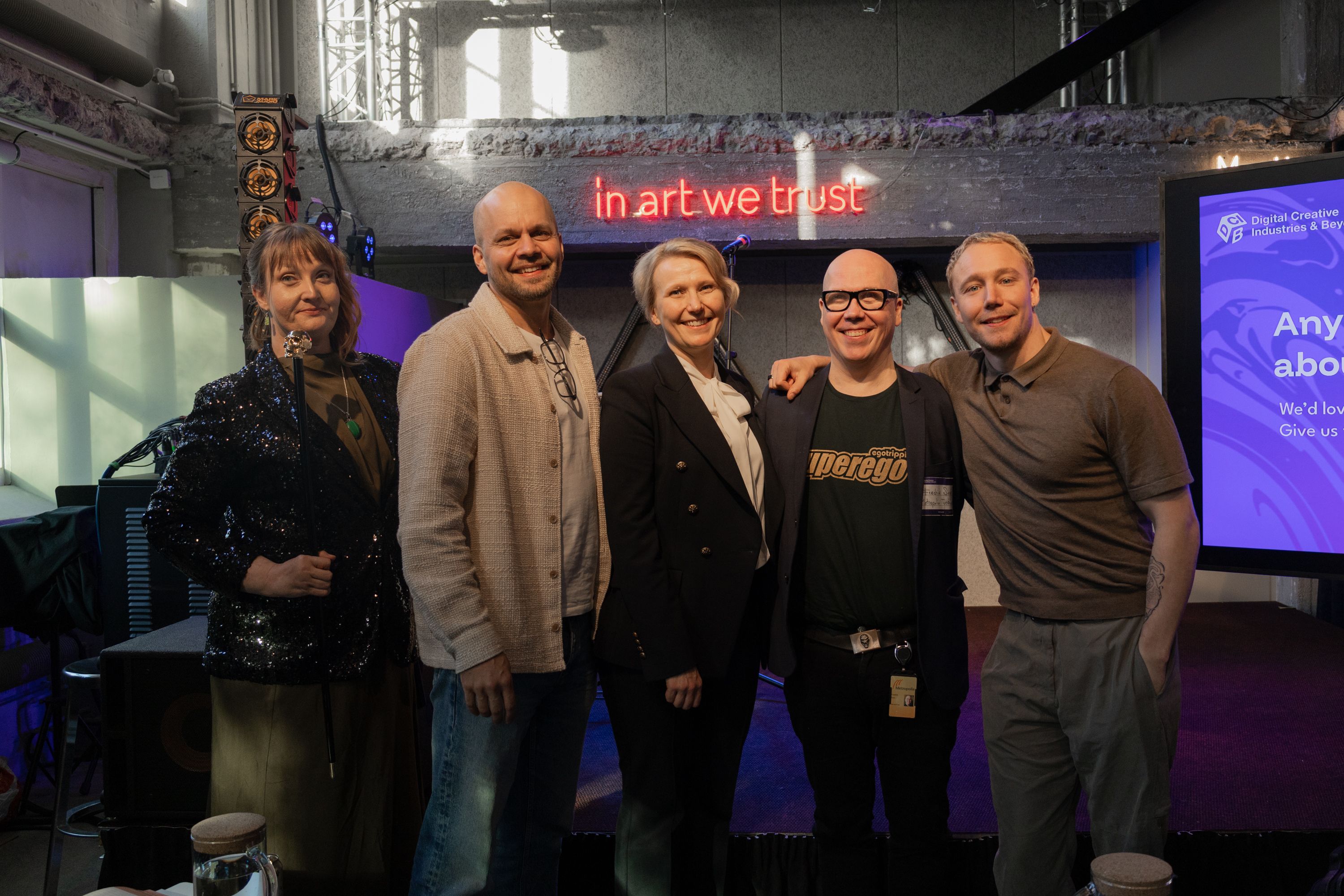 Five people standing together on a stage under a red neon sign that reads in art we trust, Ronny Eriksson on the right