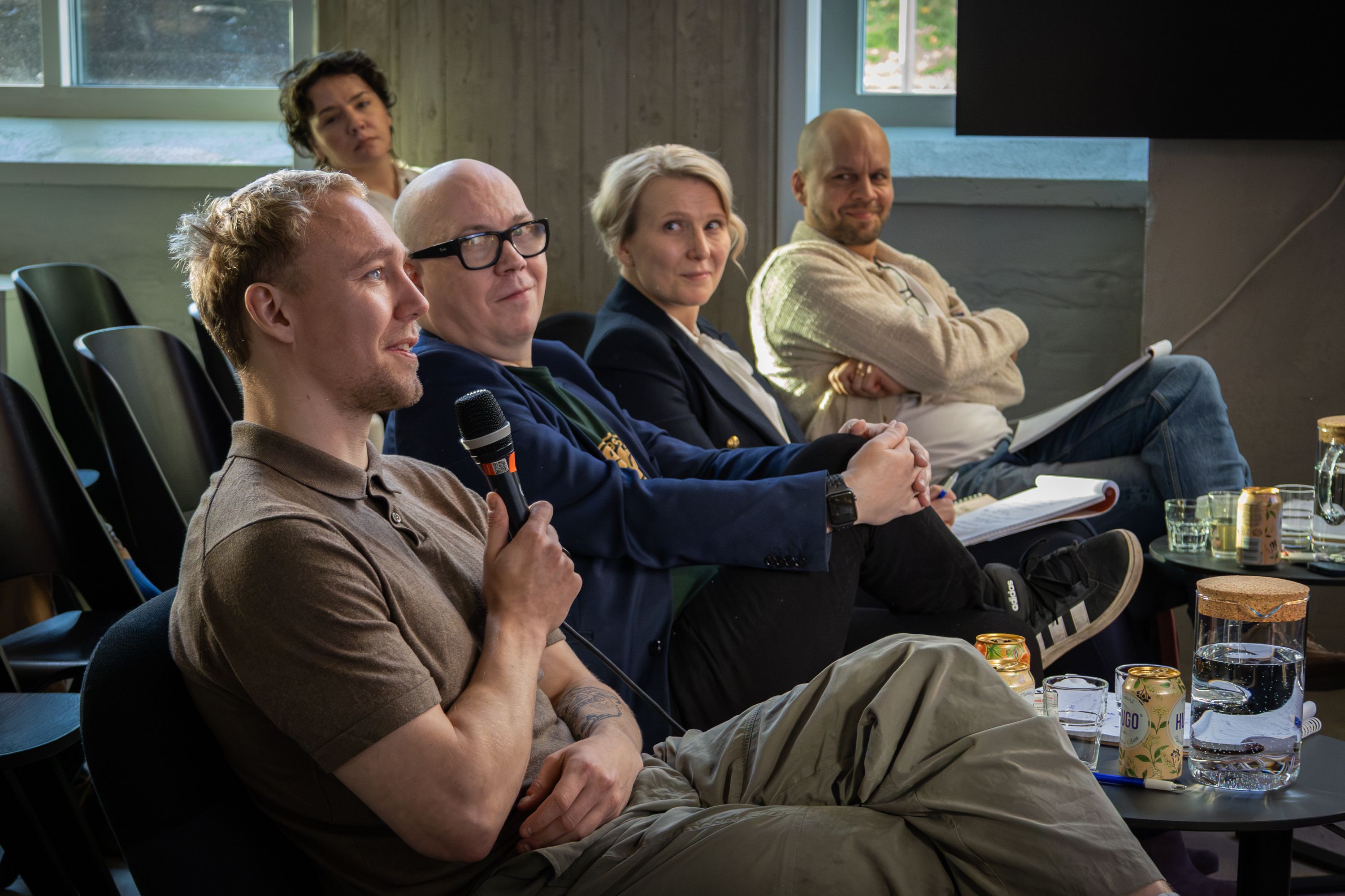 Ronny Eriksson laughing, a microphone in his hand, at the end of a jury of four seated in a row by tall windows