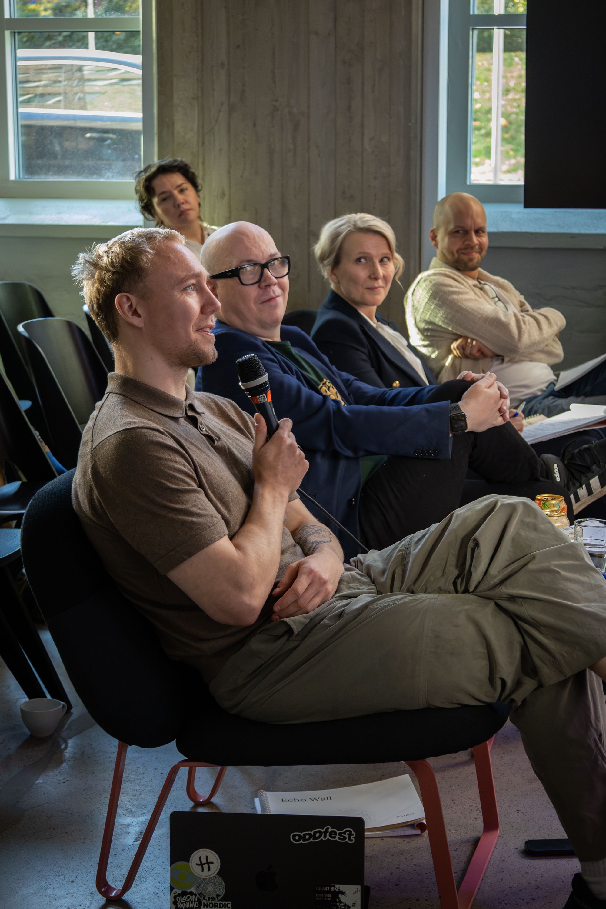 Ronny Eriksson in a brown polo shirt with a microphone, smiling, the rest of the jury seated beside him