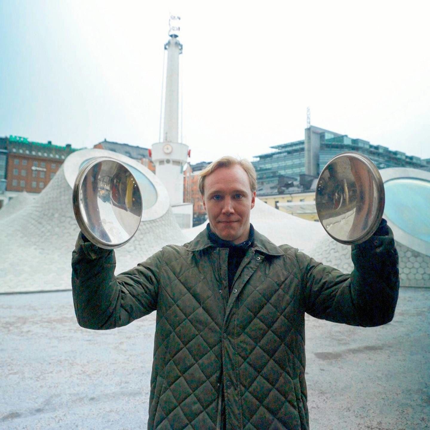 Ronny Eriksson holding two convex mirrors up at shoulder height on Lasipalatsi square in Helsinki