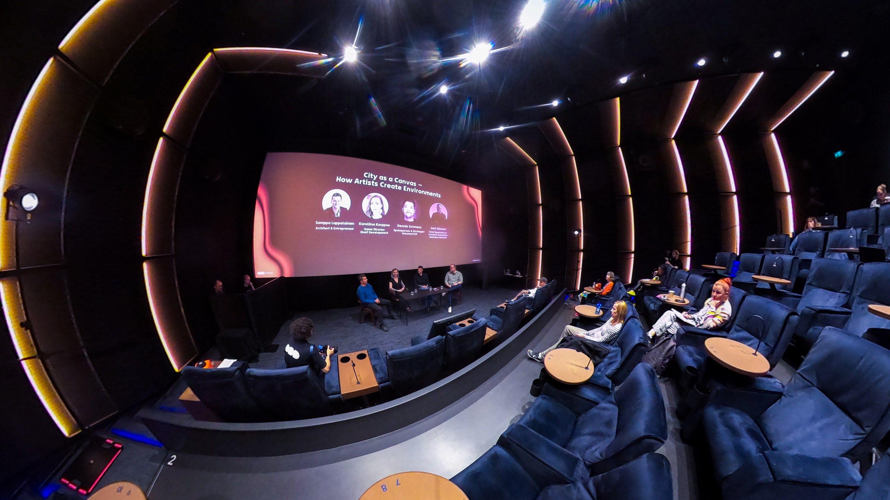A wide-angle view of a cinema auditorium lined with blue armchairs, a panel on stage under a pink screen