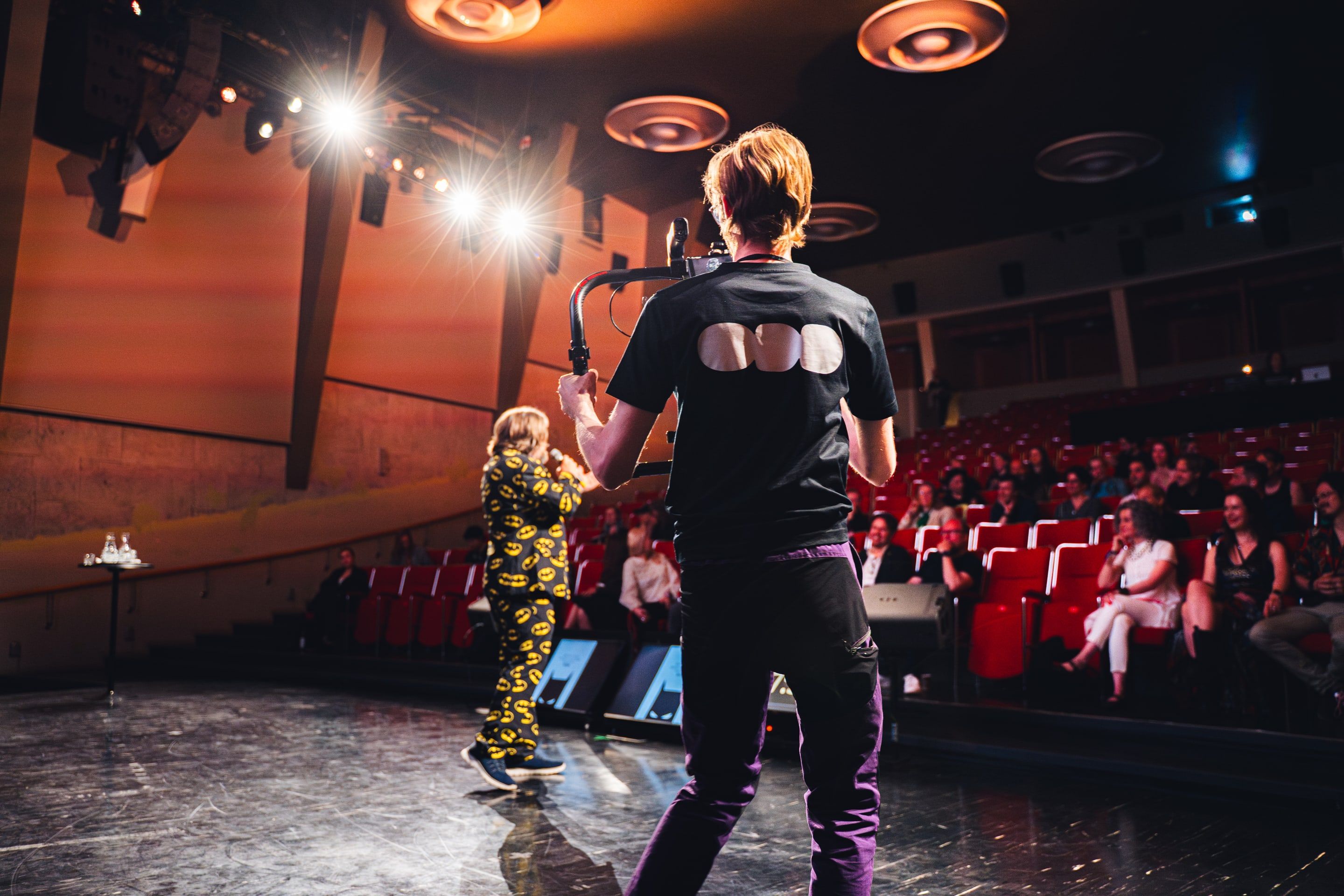 A camera operator in an ODD t-shirt filming Claus Raasted in a Batman-print suit on the Bio Rex stage, a laughing audience in red seats