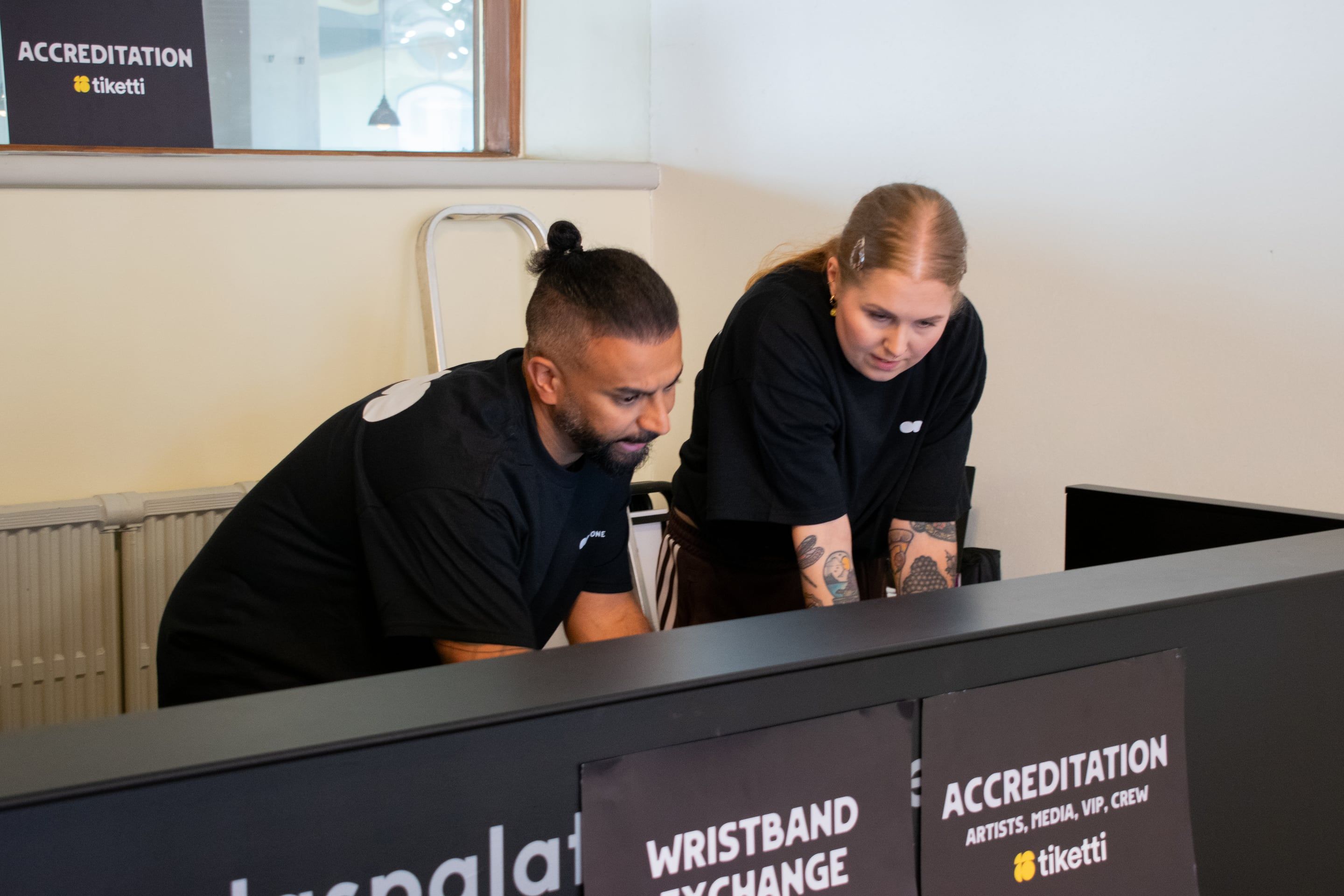 Two volunteers in ODD t-shirts leaning over the accreditation and wristband desk together, working something out on a screen