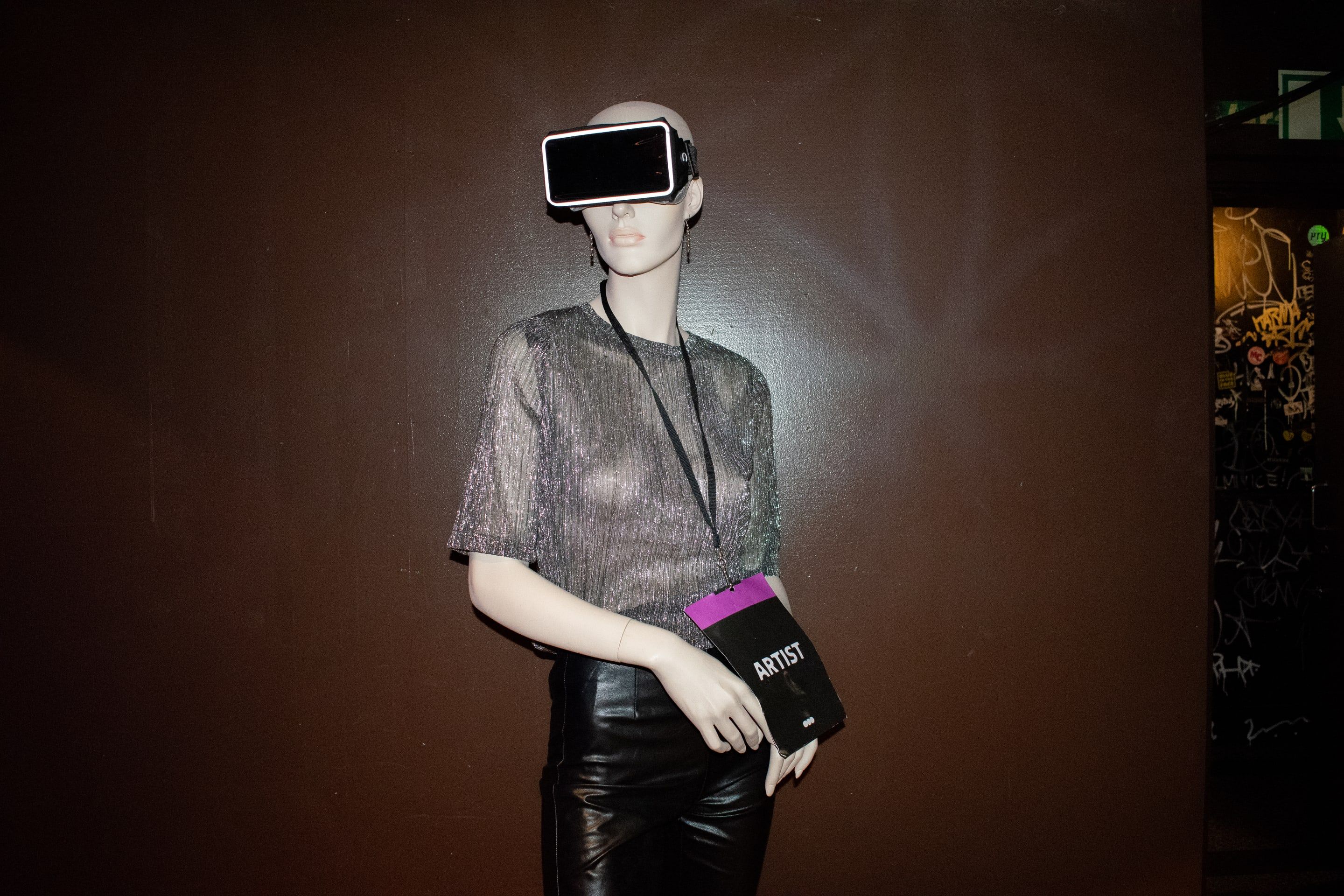 A mannequin in a silver top and a VR headset holding an artist pass, against a brown wall