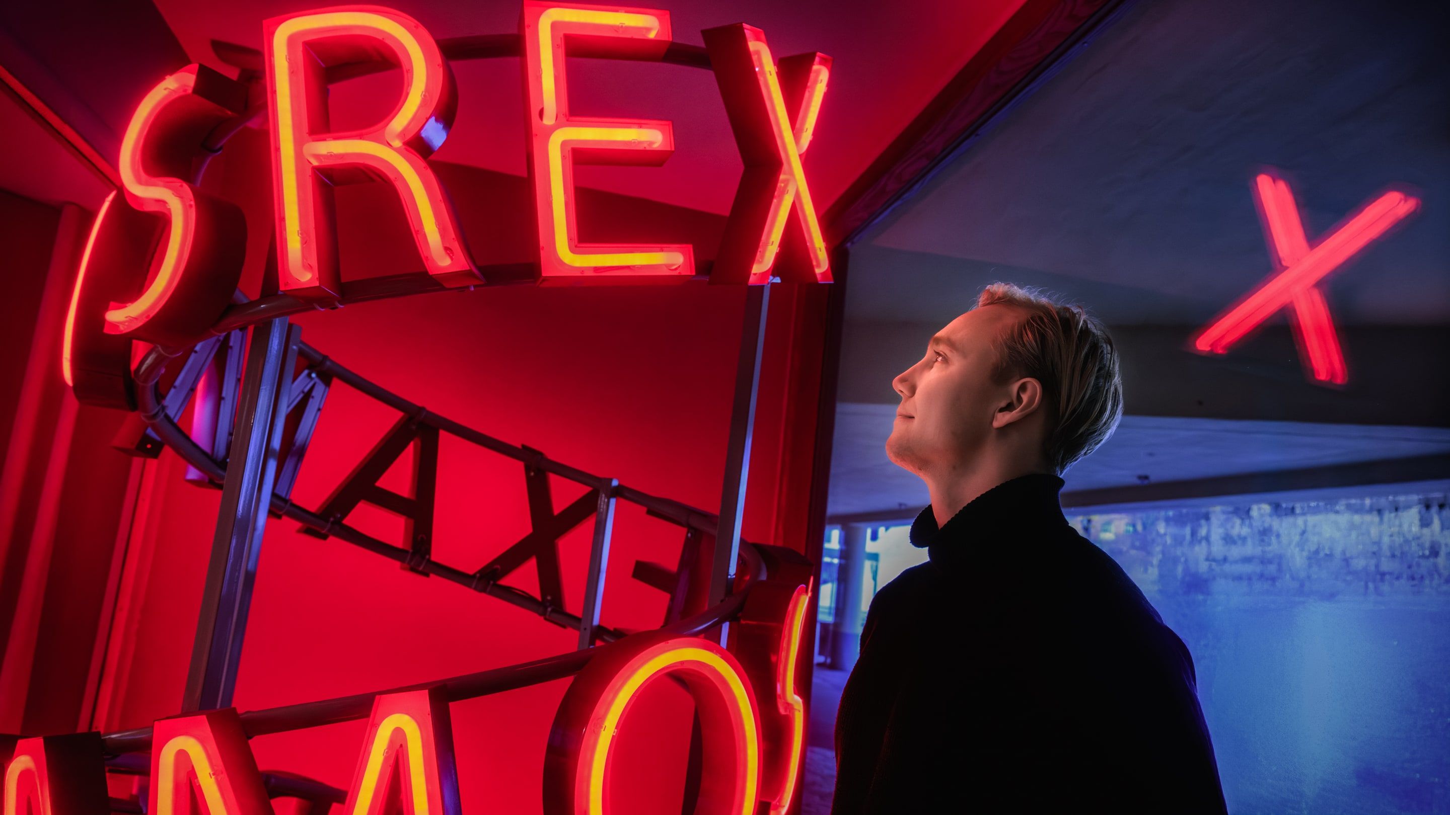 Ronny Eriksson in profile on the right, looking up at large red neon letters on the left, cool blue space around his head