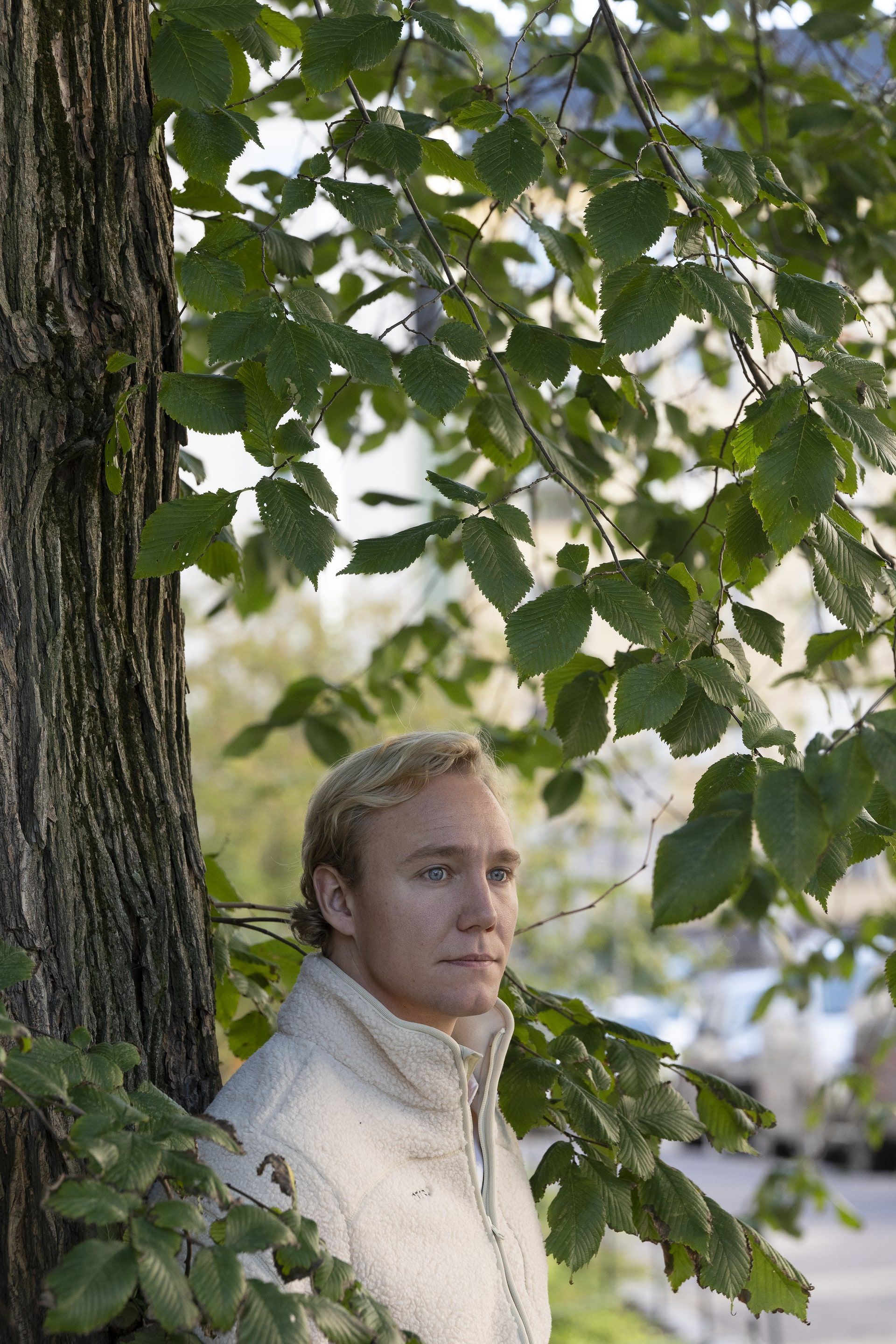 Ronny Eriksson in a cream fleece leaning beside a tree trunk, green leaves hanging above him