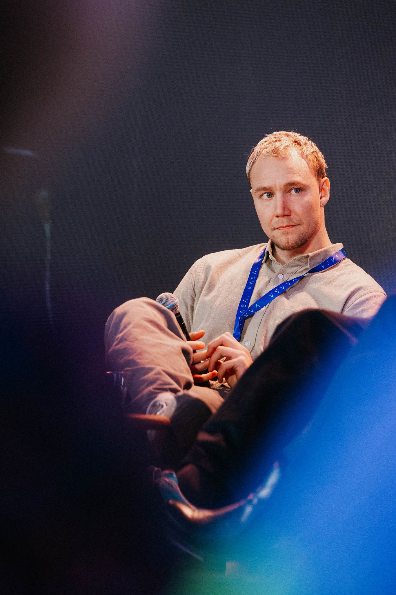 Ronny Eriksson in a beige jumper in an armchair on stage, legs crossed, microphone in hand, listening to another panellist