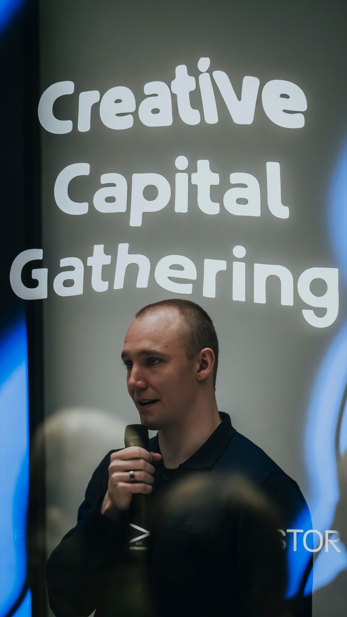 A speaker with a microphone under a screen reading Creative Capital Gathering