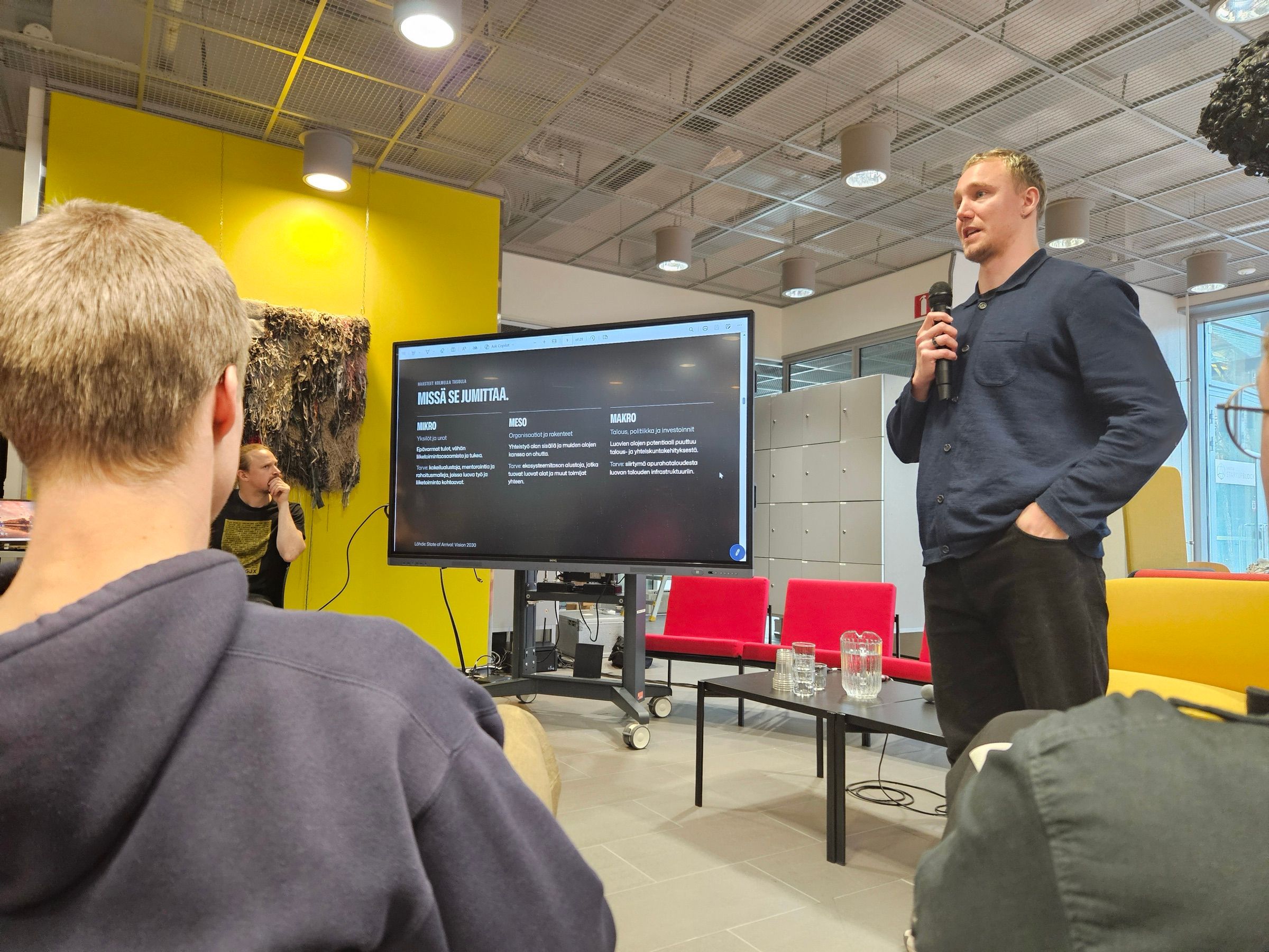 Ronny Eriksson with a microphone beside a screen that reads Missä se jumittaa., red and yellow chairs behind him
