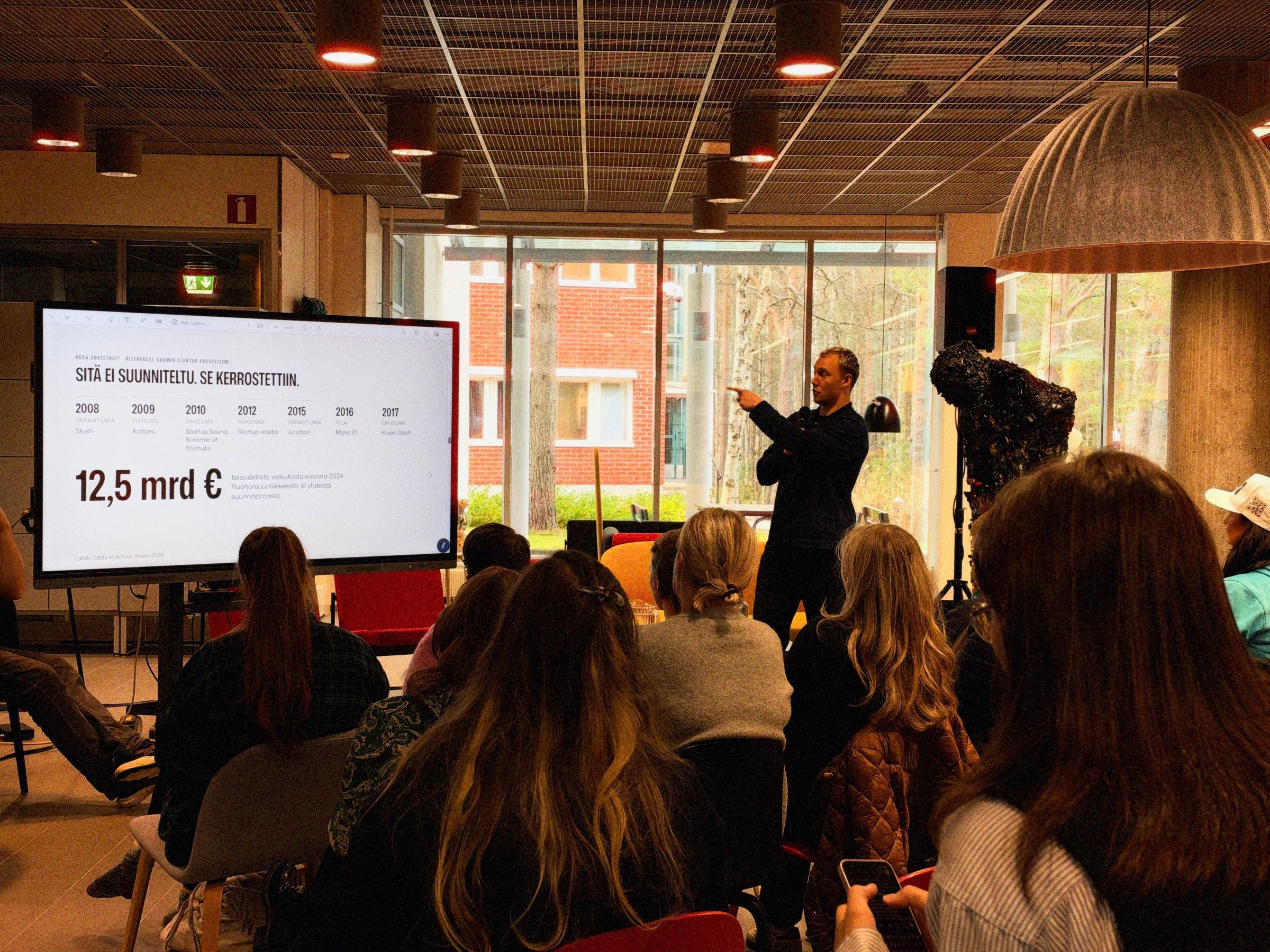 Ronny Eriksson pointing at a screen that reads Sitä ei suunniteltu. Se kerrostettiin., the F-hub audience seen from behind, birches outside the window