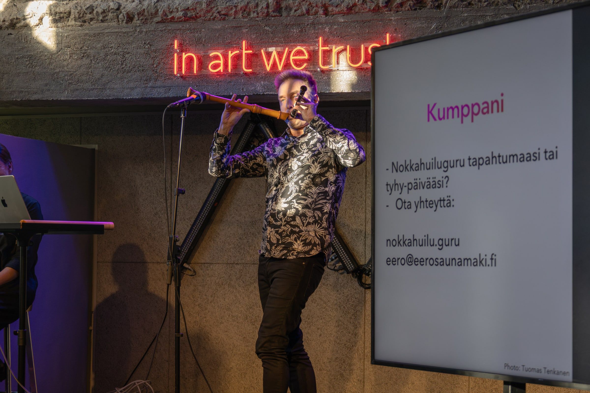 Eero Saunamäki i mönstrad skjorta spelar en lång blockflöjt under neonskylten, bredvid honom en bild där det står Kumppani