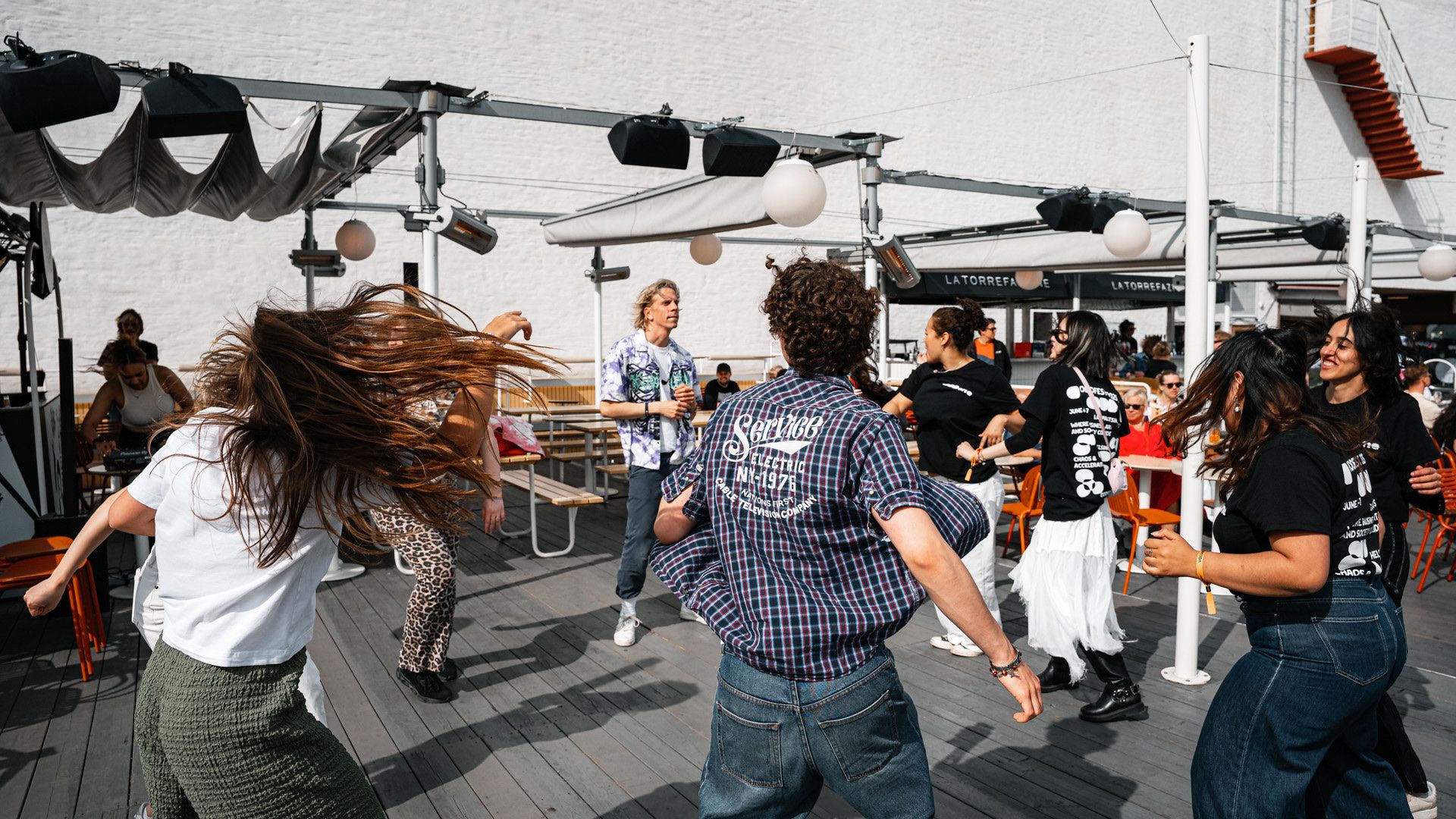 Dancers pulling a crowd in shirts and lanyards into a dance under a canopy, in daylight